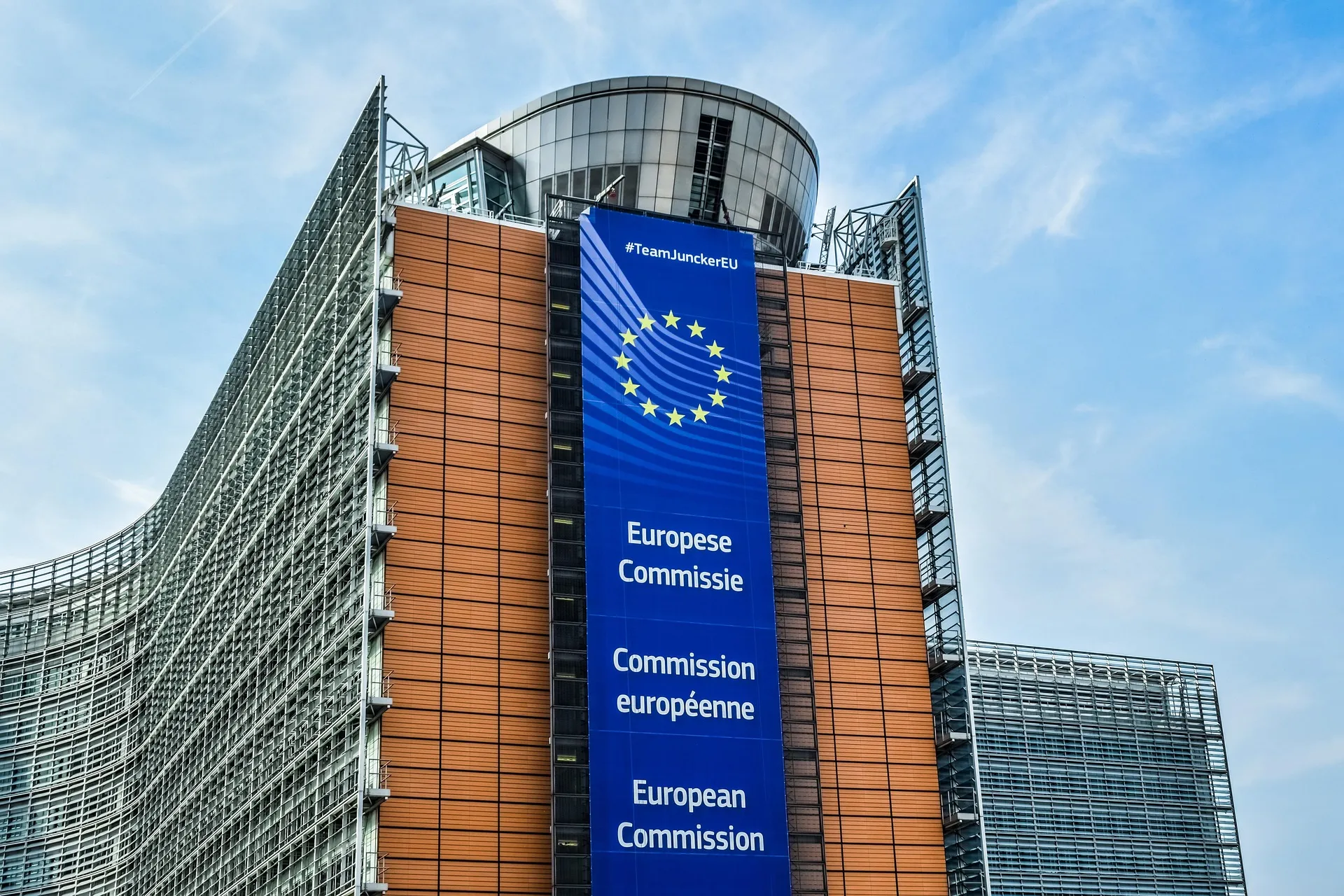 The European Commission headquarters in Brussels seen from below, a tall banner down its facade carrying the European Union's circle of stars above the words “Europese Commissie”, “Commission européenne” and “European Commission”.