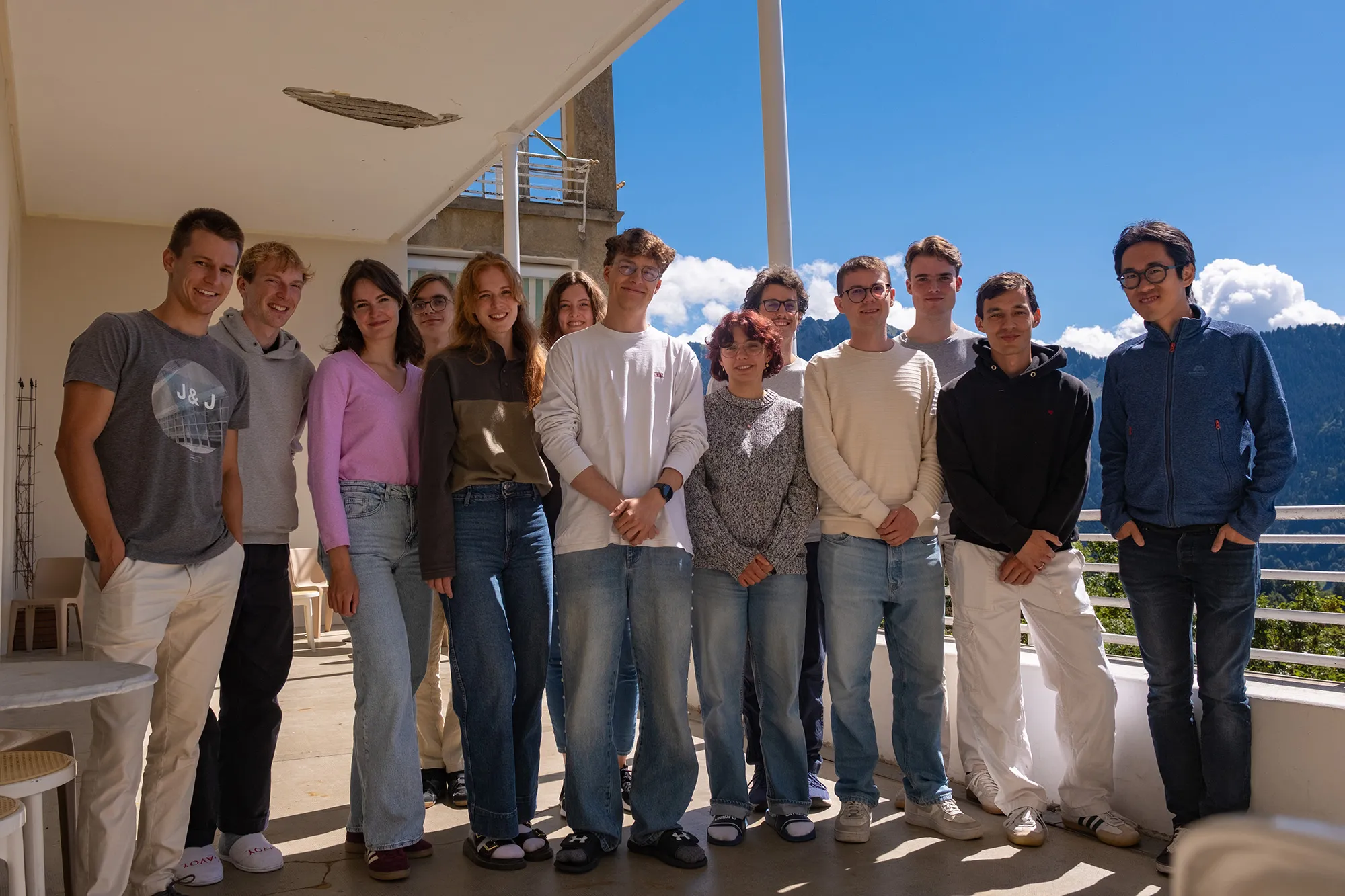 The group standing side by side on a covered terrace, with a wooded mountain slope and a blue sky behind them.