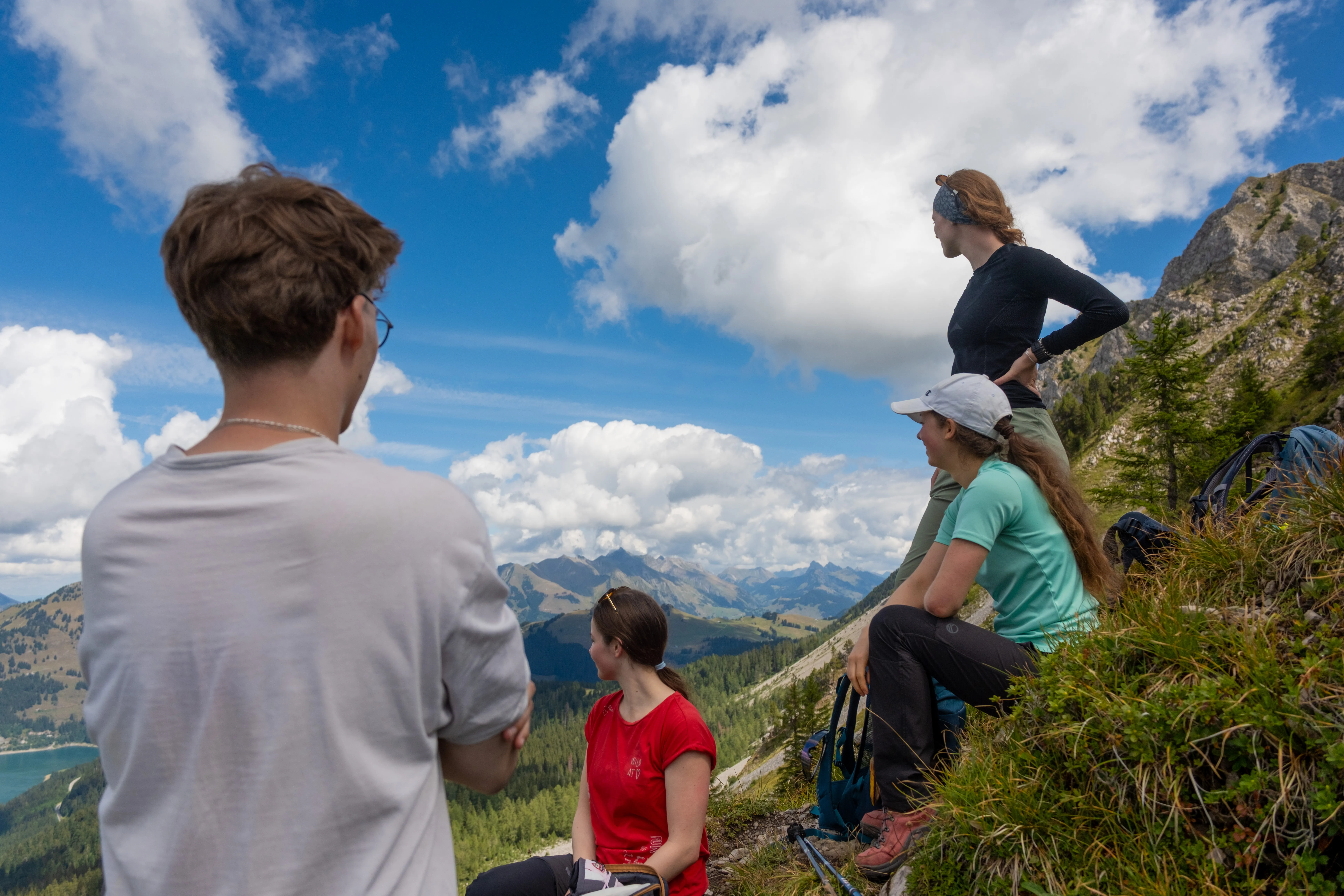 Four young hikers resting on a grassy ridge in the Swiss Alps, with rucksacks beside them, a lake below and mountain ranges under a partly clouded sky behind.